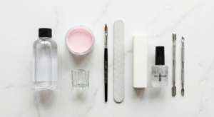 Flatlay of essential manicure tools including monomer, powder, brush, and file on white marble