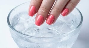 Freshly painted coral pink nails being dipped into a bowl of ice water with visible condensation for quick drying.