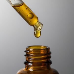 A golden droplet of facial serum hanging from a glass pipette above a dark amber bottle.