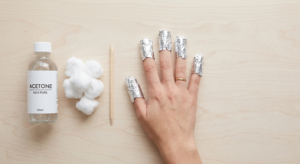A top-down view of the foil-wrap method for safe gel polish removal, showing a hand with foil-wrapped fingertips next to acetone, cotton balls, and a cuticle stick.